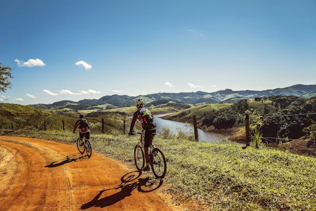 How to Track Outdoor Cycling with Apple Watch 5 Two cyclists riding mountain bikes along a winding dirt road in a scenic countryside with hills and a river in the background.